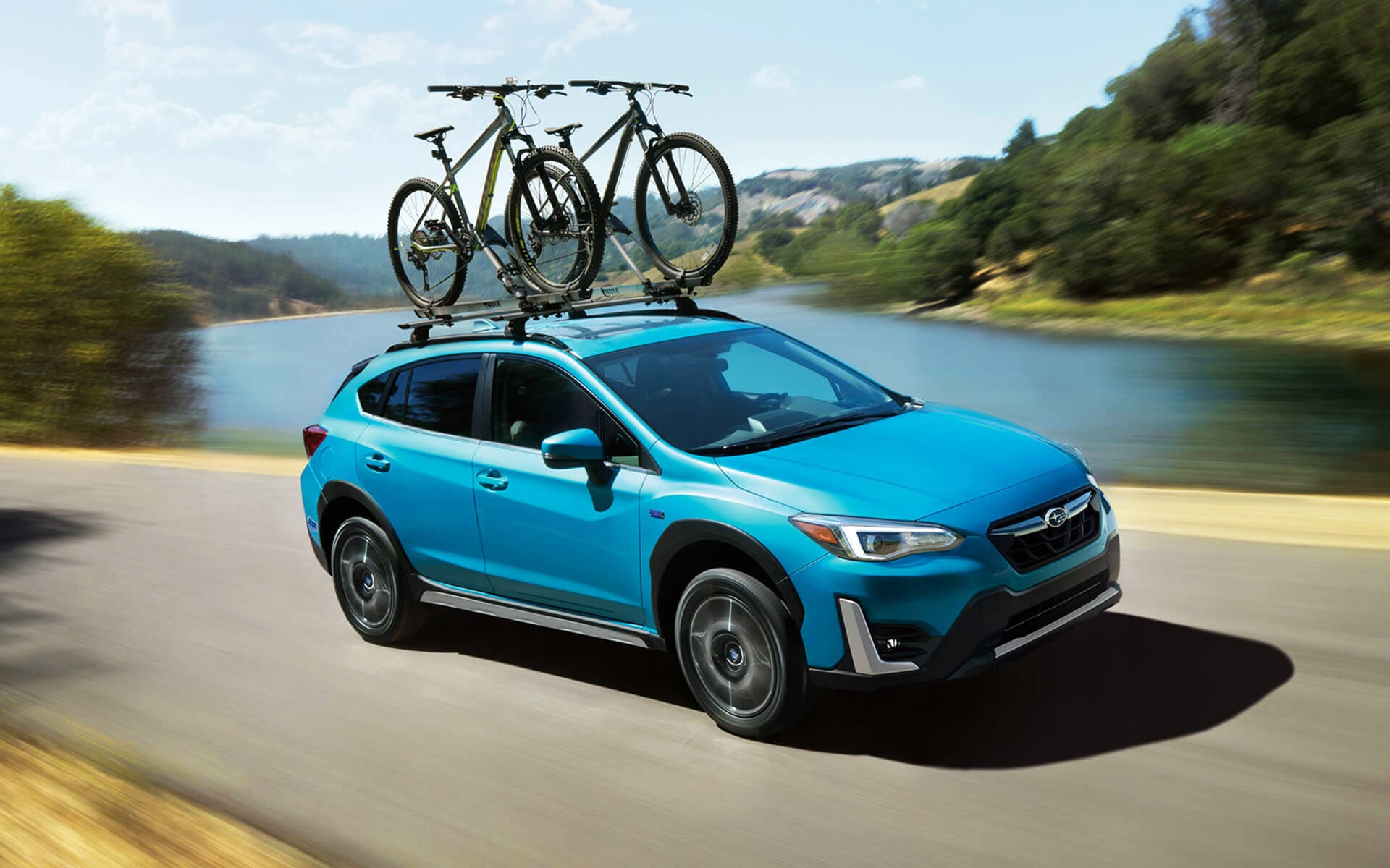 A blue Crosstrek Hybrid with two bicycles on its roof rack driving beside a river | Island Subaru in Staten Island NY